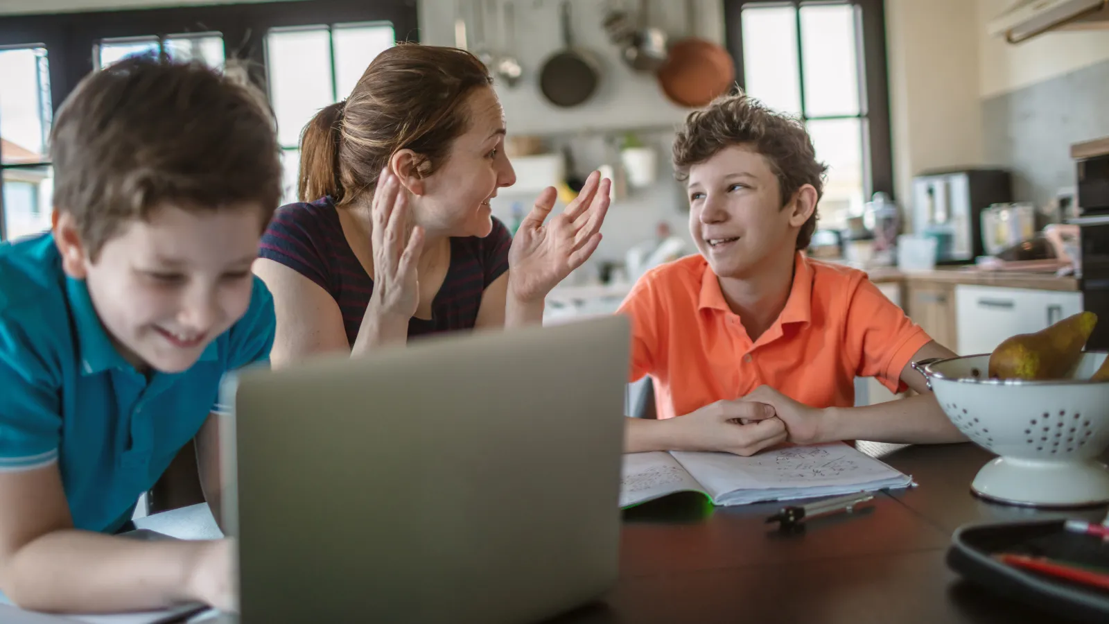 Younger boy working on computer with mother explaining to another child in a kitchen.